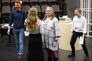 Three students and a director rehearse a scene of a play. The director talks to one students as another two laugh in the background. 