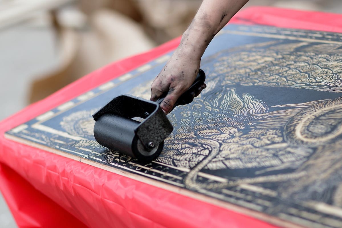 A large printing block is inked using a roller.