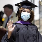 Person in grad regalia and mask waves to camera