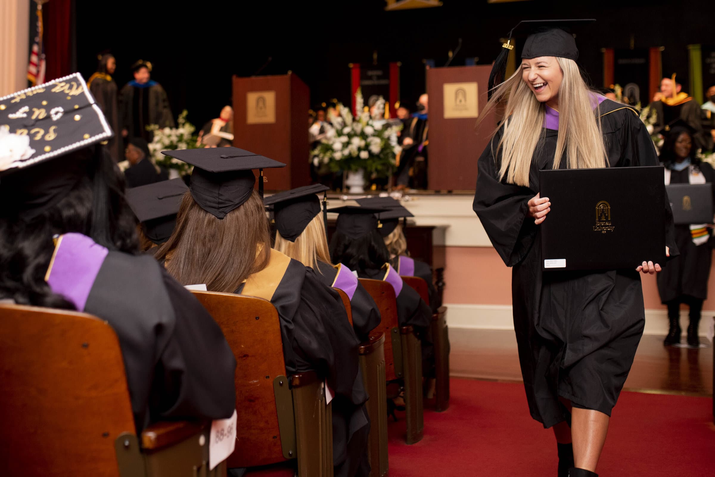 A graduate student smiles at her fellow graduates as she returns to her seat with her diploma.