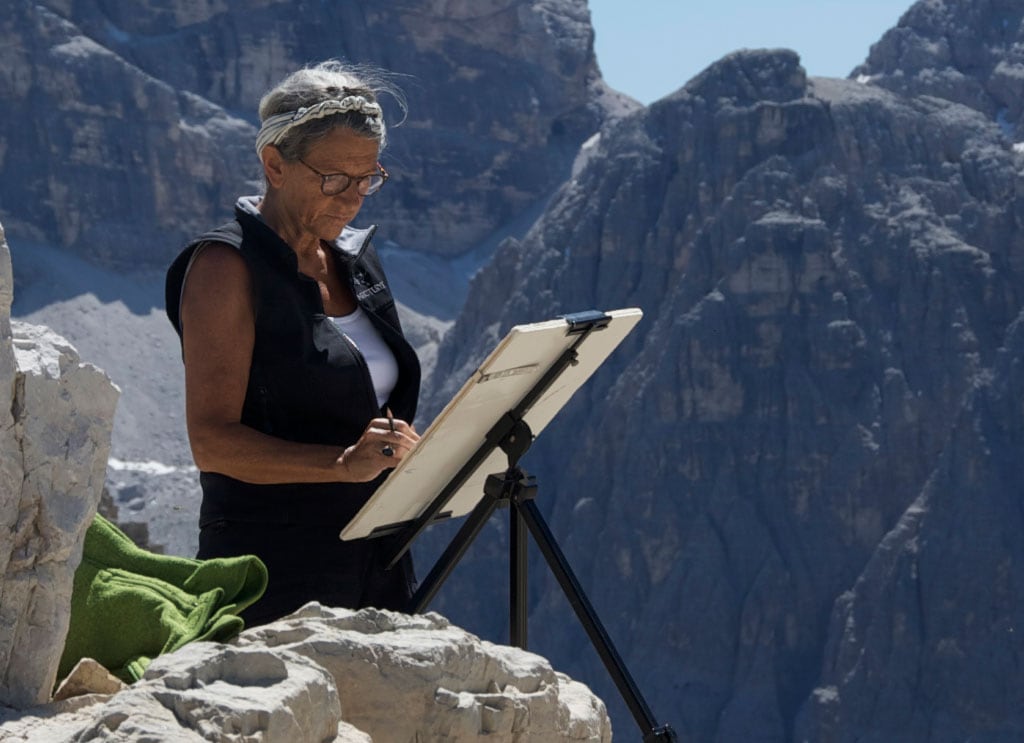 Riccarda de Eccher paints at a summit with mountains behind her