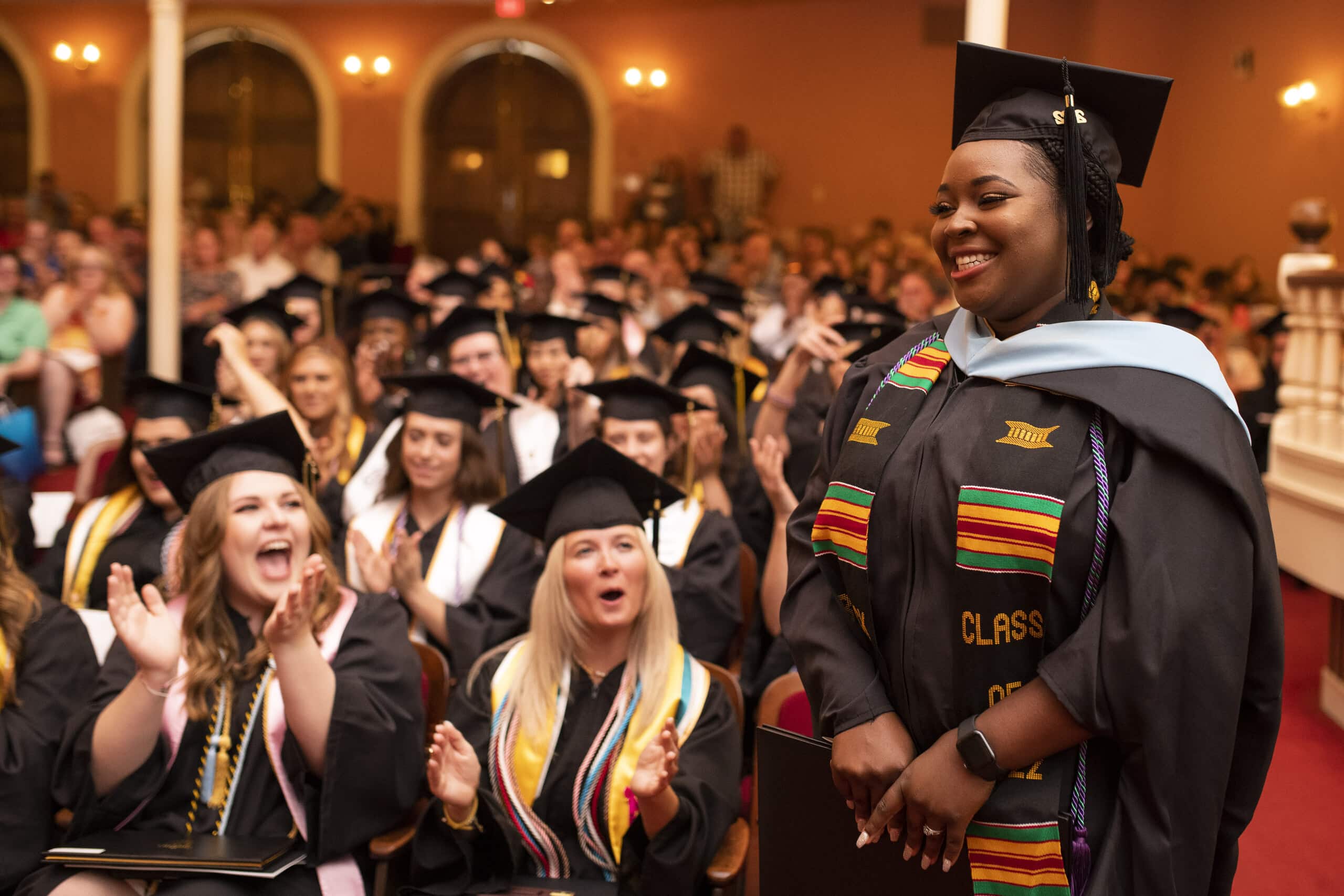 Seated audience of young women in graduation attire applaud a standing young women in graduation robes