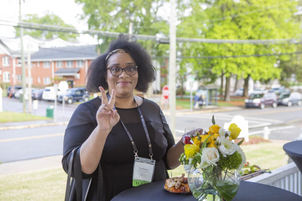 alumna stands at a table, gives the peace sign