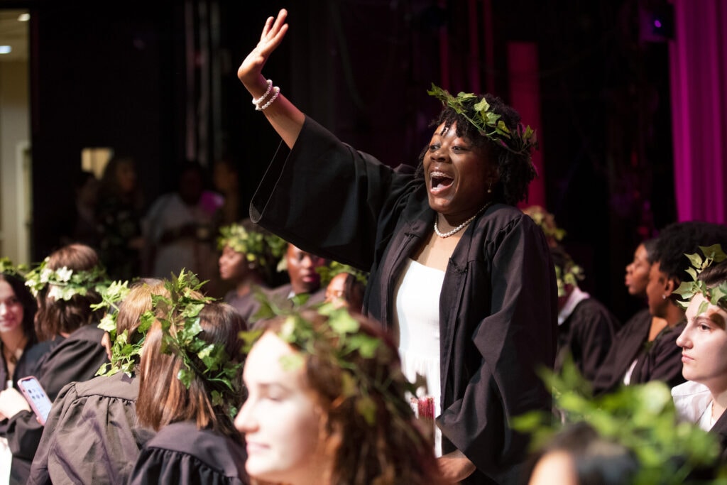 woman in black robe with ivy crown stands and waves