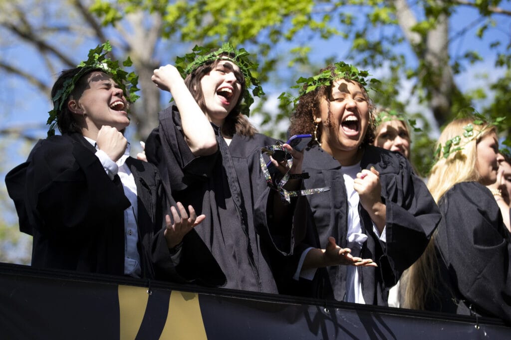 young women in black robes wearing ivy crowns wave