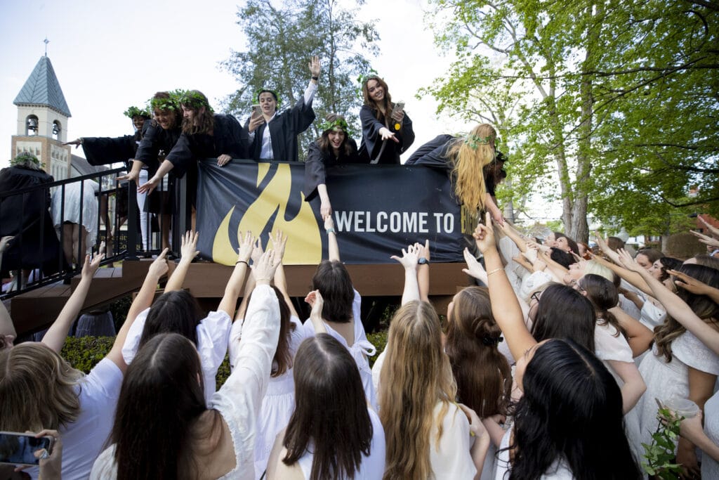 group of women in white dresses reaches up to group of women in black robes