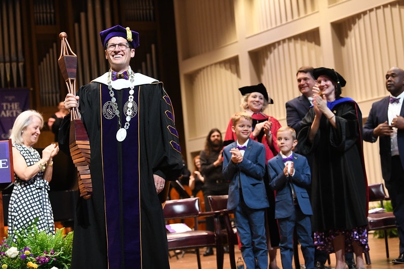 A university president wearing academic regalia holds a ceremonial mace as others behind him applaud.