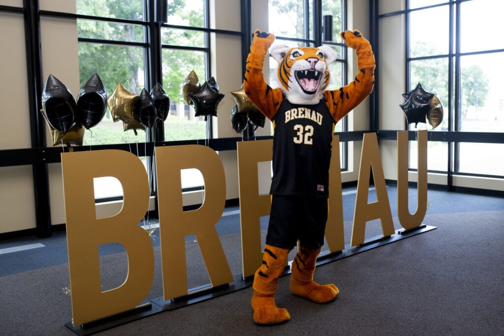 HJ the golden tiger mascot poses, paws up, in front of letter cutouts that spell Brenau.