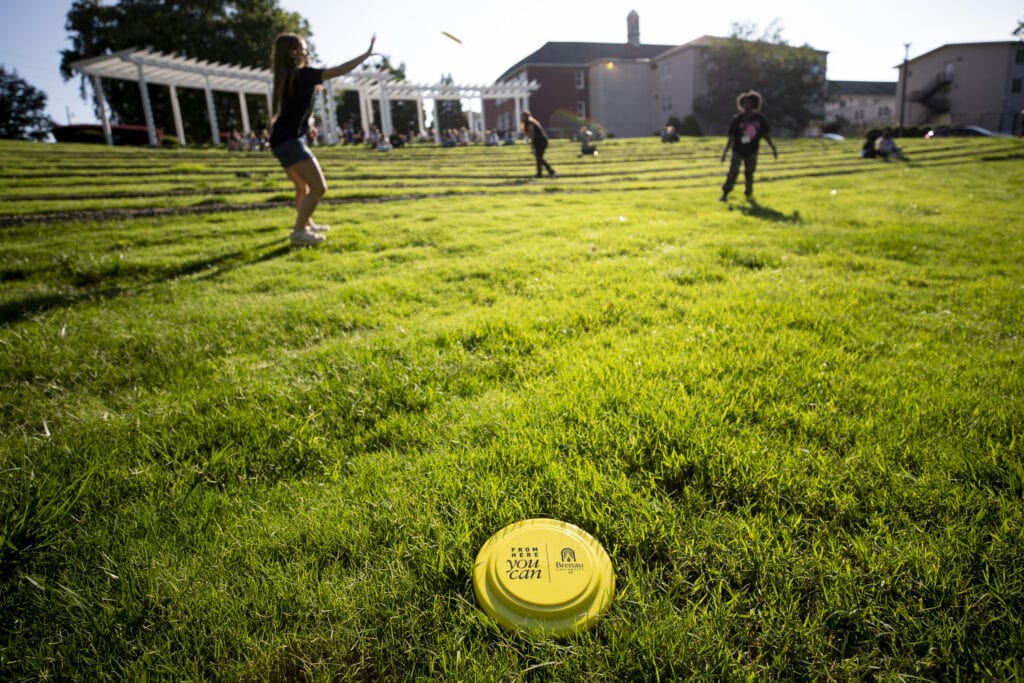 A yellow Brenau frisbee lays in the grass of the amphitheatre, with students playing in the background.