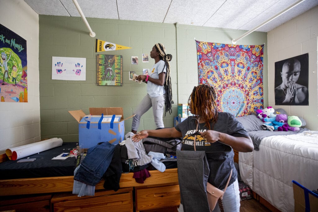 A student stands on her lofted bed as she hangs a poster; her mother organizes her clothes.