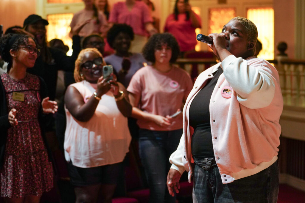 India Williams sings into a microphone while a student films her on a phone