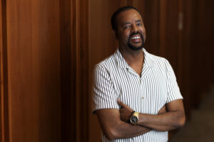 A man in a short sleeve shirt stands in front of a wood-paneled wall.