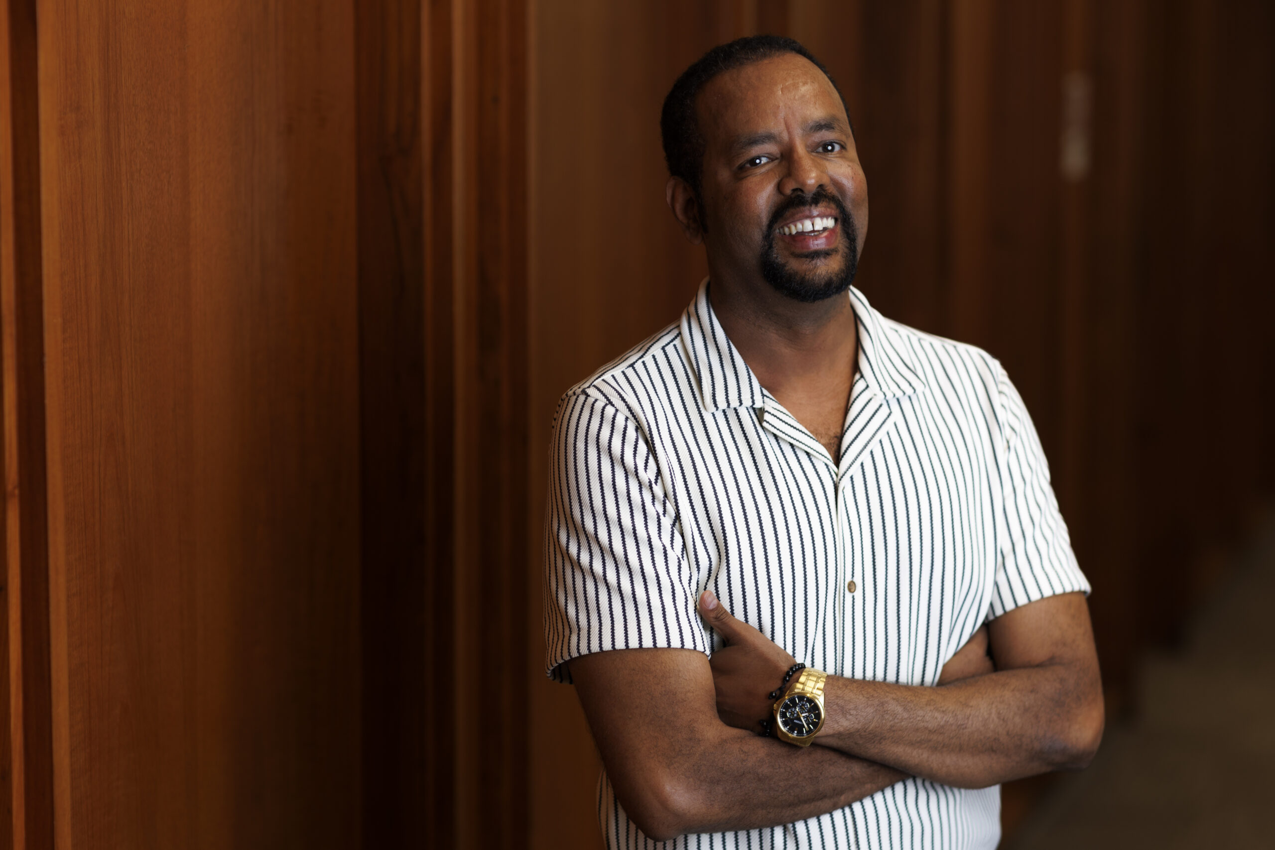 A man in a short sleeve shirt stands in front of a wood-paneled wall.