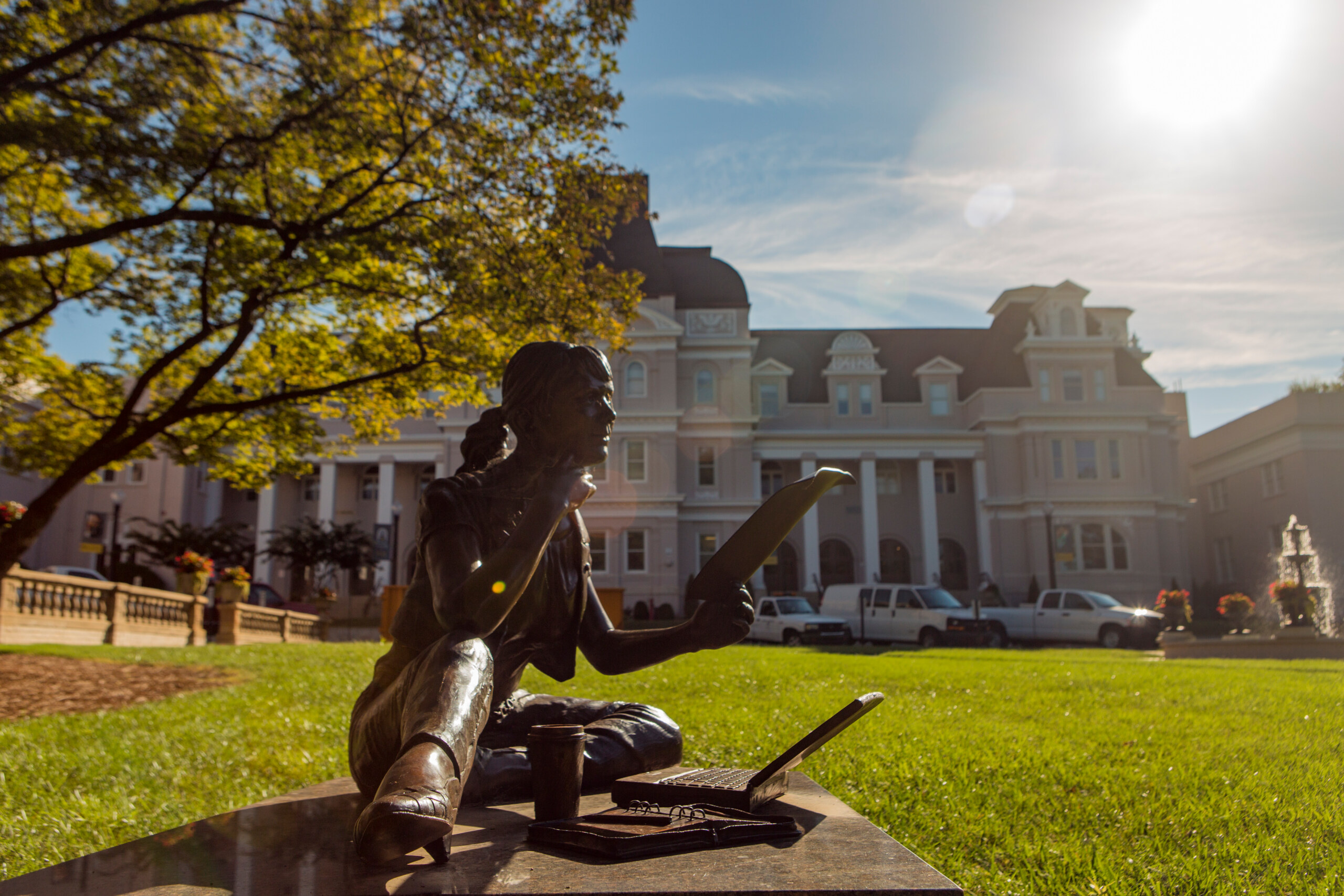 bronze status of a young woman seated on the lawn in front of a university building.