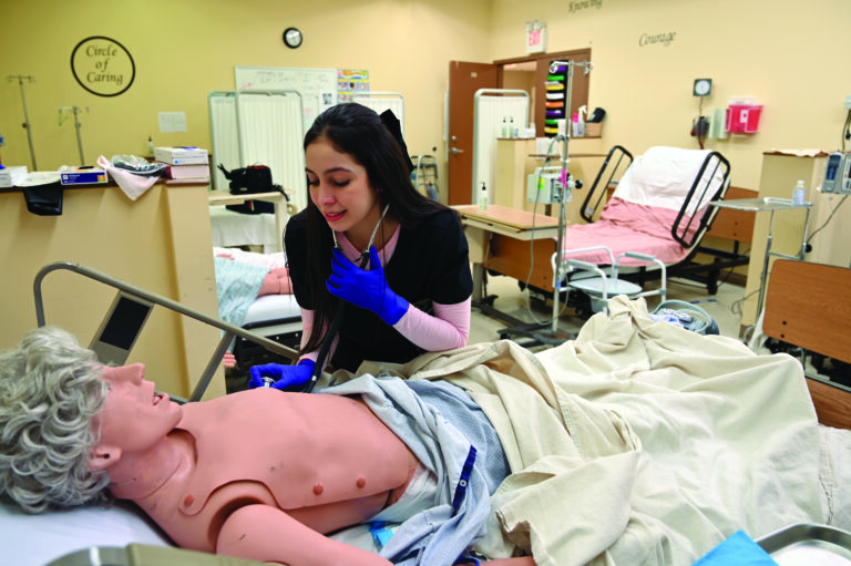 Laura Trimboli, a nursing student, holds a stethoscope close to a dummy laying on a hospital bed.