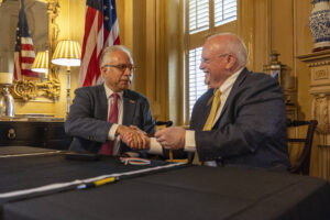 Two men in business suits shake hands while seated at a table.