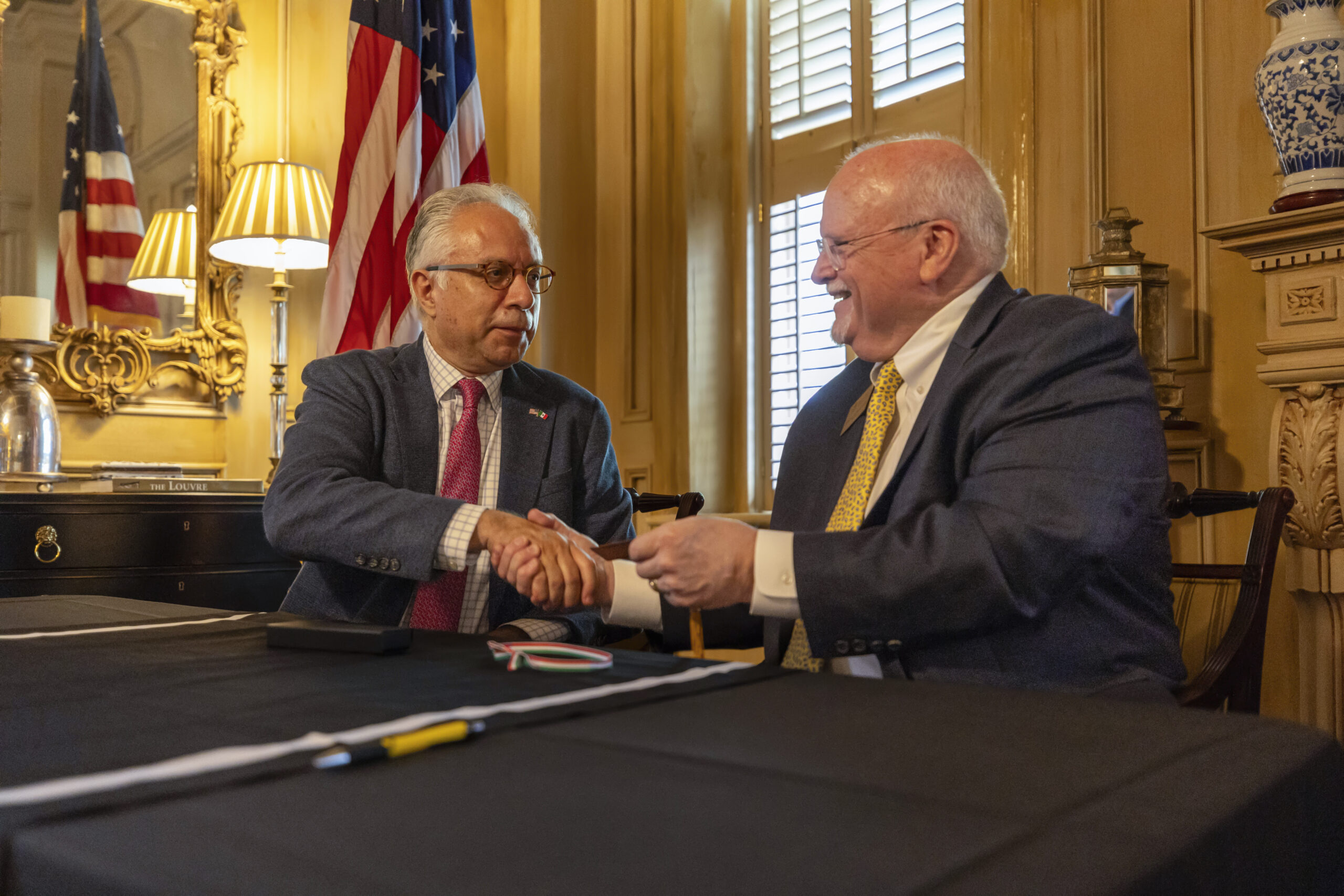 Two men in business suits shake hands while seated at a table.