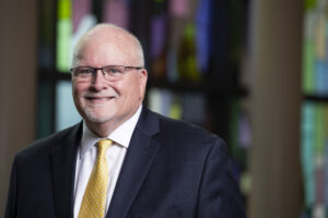 Brenau President David L. Barnett in a dark suit and gold tie stands in front of a stained glass window.
