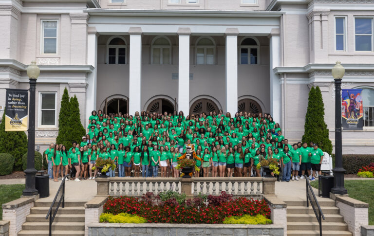 large group of students wearing green shirts stands on the steps of a traditional college building
