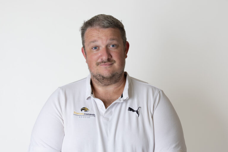 Zac Reichert wearing a white polo shirt stands in front of white background.