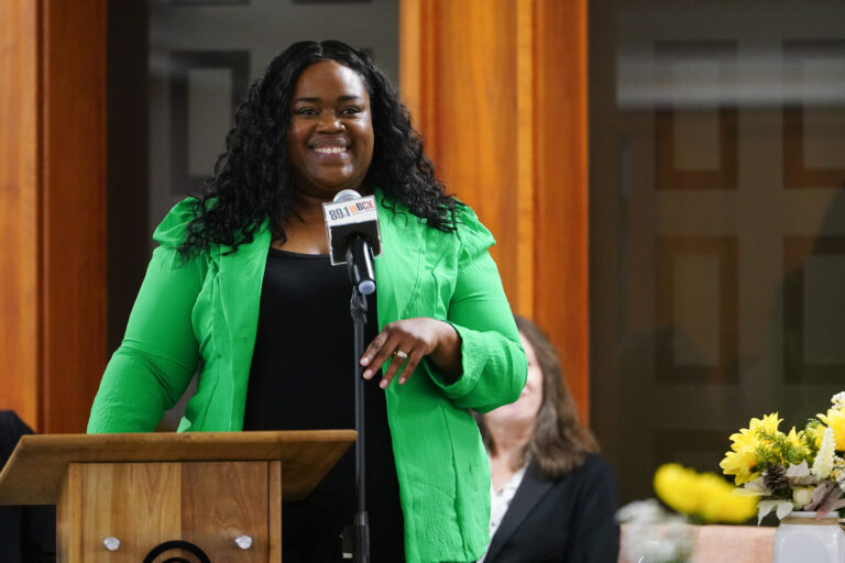 Shatrela Washington-Hubbard wearing a black blouse and green blazer smiles while holding a microphone.